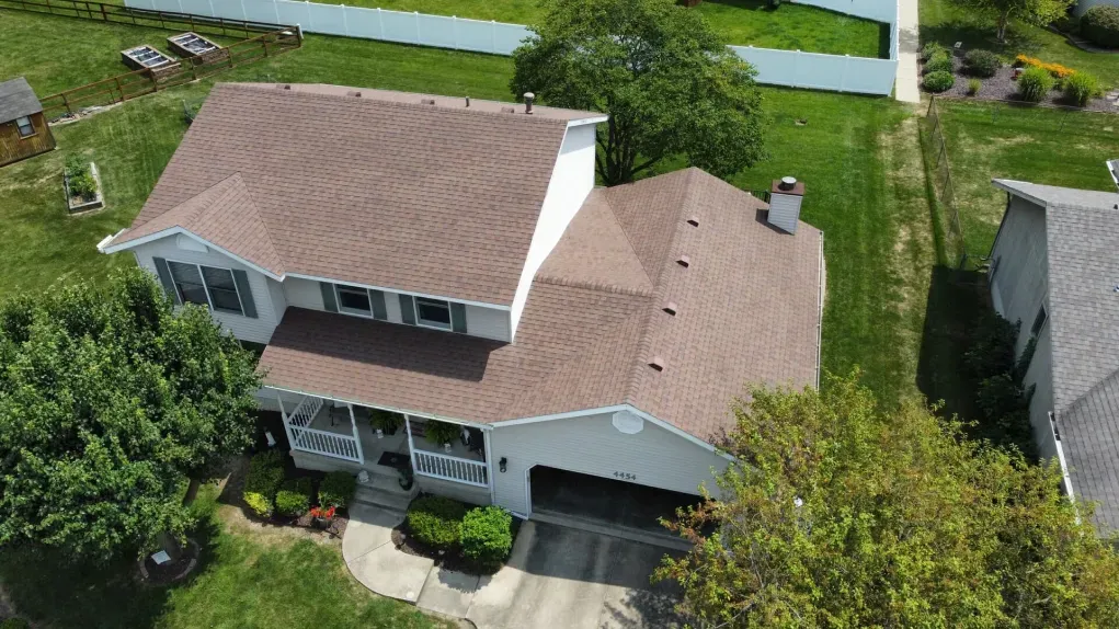 An aerial view of a two-story house with a brown shingled roof, a paved driveway, and surrounding green lawn and trees.