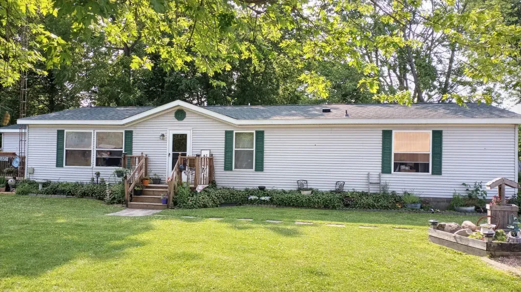 A light gray manufactured home with green shutters and a front porch, set in a grassy yard surrounded by trees.