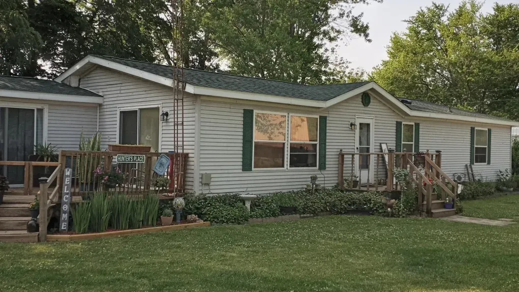 A single-story, light-colored manufactured home with green shutters and a wooden deck set against a grassy, tree-lined lawn.