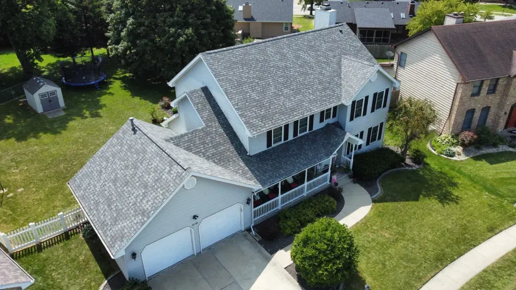 An aerial view of a two-story light gray house with a gray shingled roof, a front porch, and a driveway in a suburban yard.