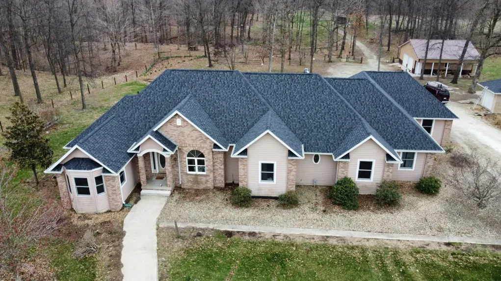 An aerial view of a single-story brick house with a dark shingled roof, surrounded by trees and a rural landscape.