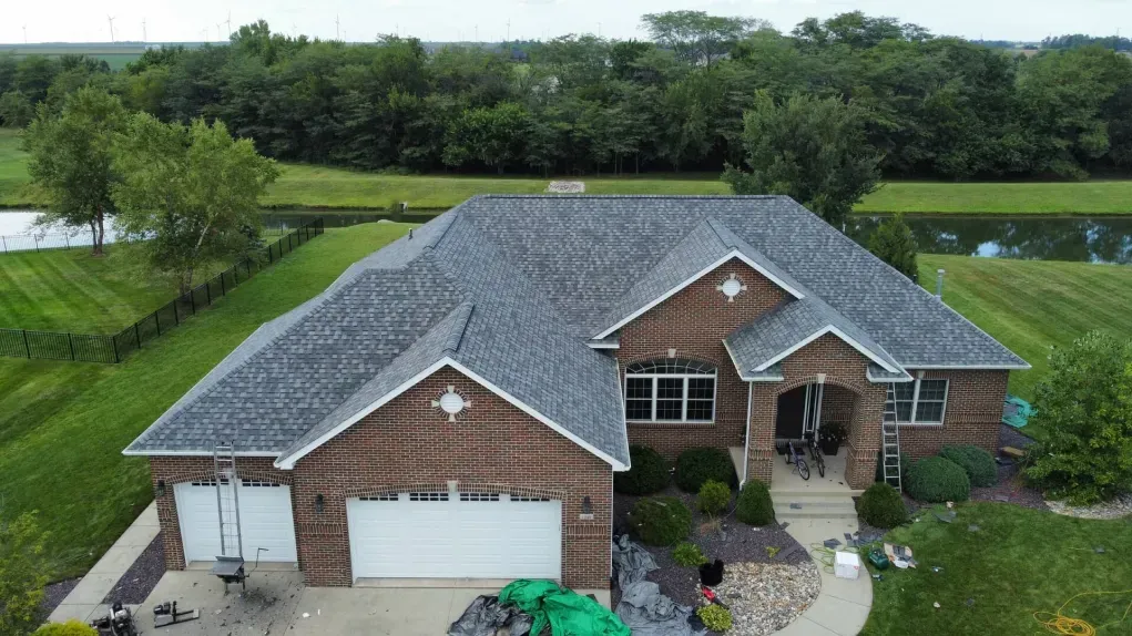 A high-angle view of a brick house with a new grey shingled roof, surrounded by a lawn and a pond in the background.