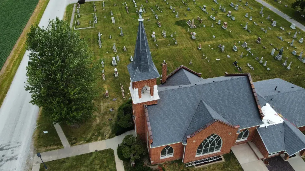 Aerial view of a brick church with a steeple, set beside a large, grassy cemetery with many headstones.