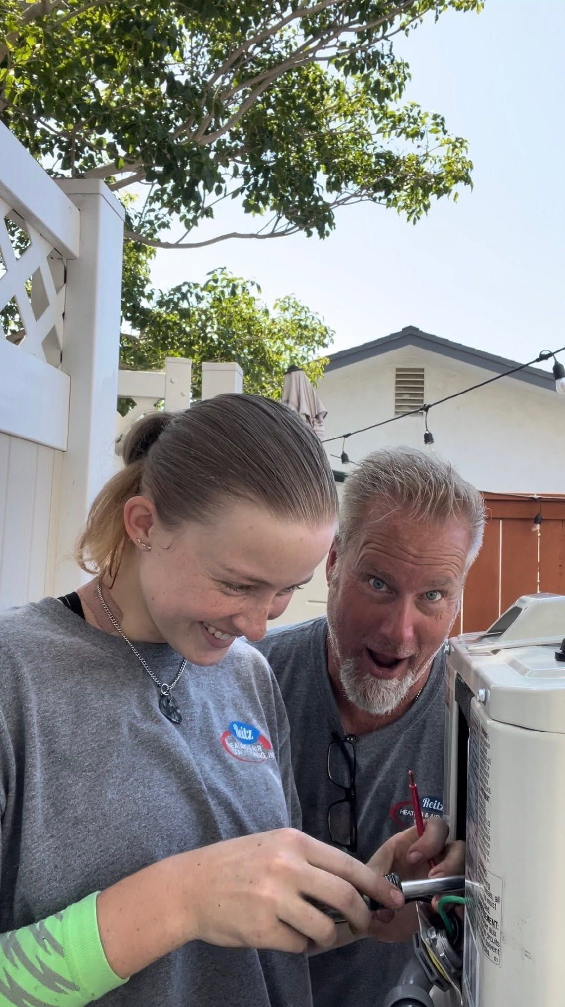 Two people smiling while using a power tool outdoors near a fence and house