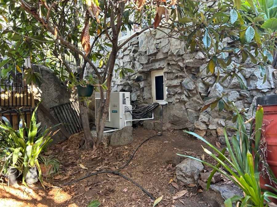 Small stone building with a doorway, surrounded by trees and plants in a yard