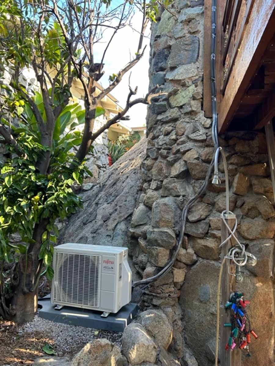 Outdoor air conditioner unit beside a stone wall, under trees and next to a wooden structure