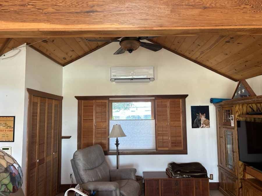 Cozy living room with wood-beamed ceiling, window shutters, recliner, lamp, and wall-mounted air conditioner