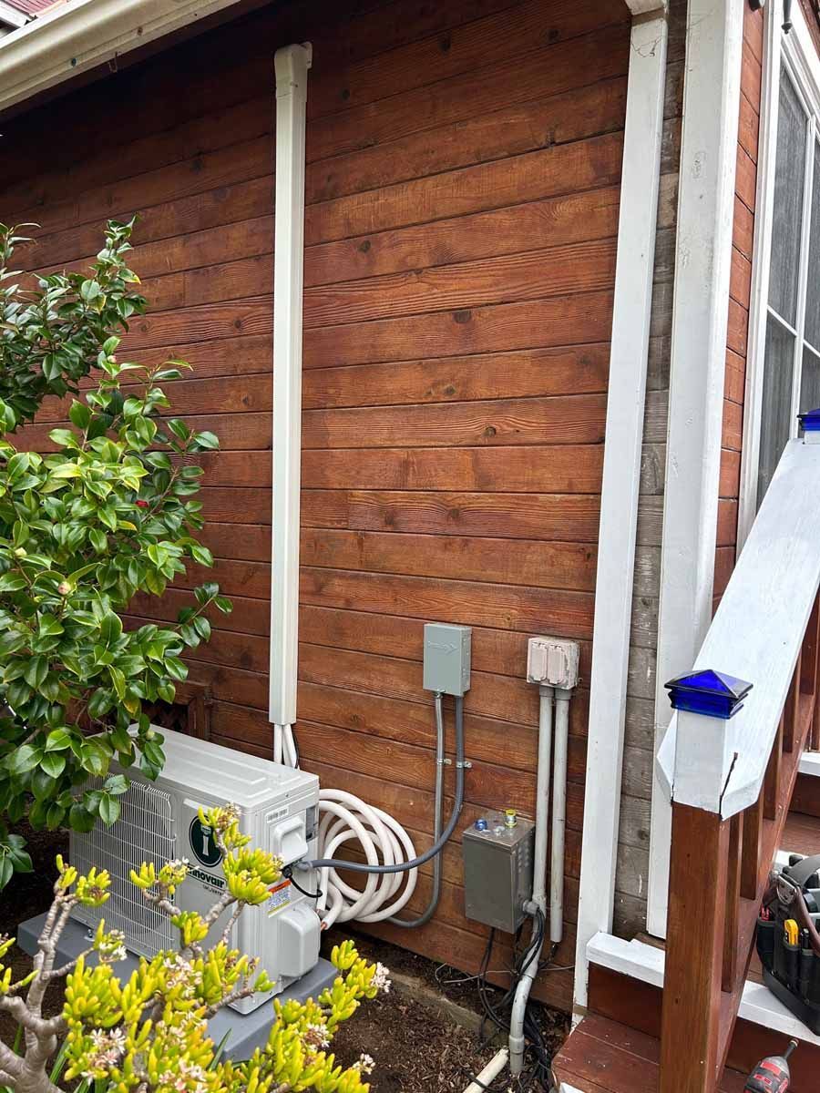 Wood-sided house exterior with utility boxes, pipes, and coiled hose beside shrubs and a hanging towel