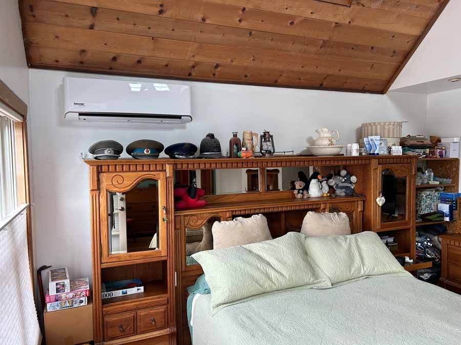 Bedroom with wooden slanted ceiling, large bed, wall-mounted AC, and shelves filled with decor and dishes