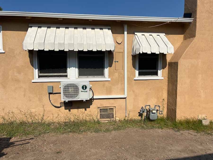 Exterior of a tan house with two windows, white awnings, and a wall-mounted air conditioner unit.