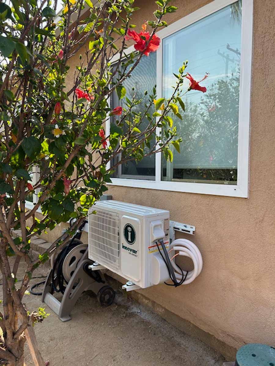 Outdoor air conditioner unit mounted on a beige wall beside a window and flowering vine