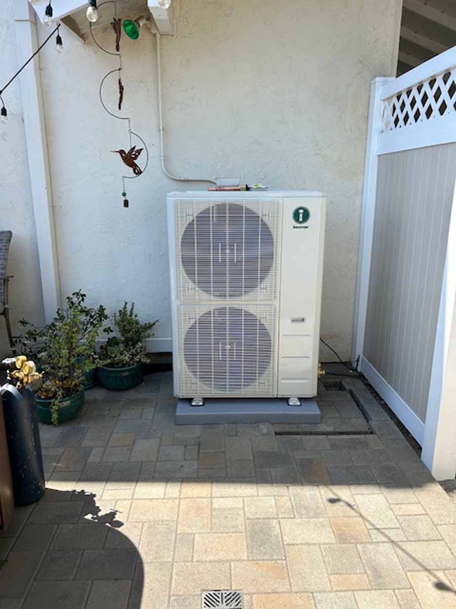 Outdoor air conditioner unit on a small paved patio beside a white wall and gate, with potted plants nearby