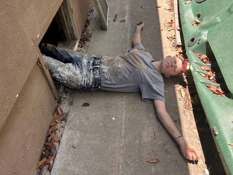 Person lying on a concrete walkway beside a building, with one arm outstretched and a green railing nearby.