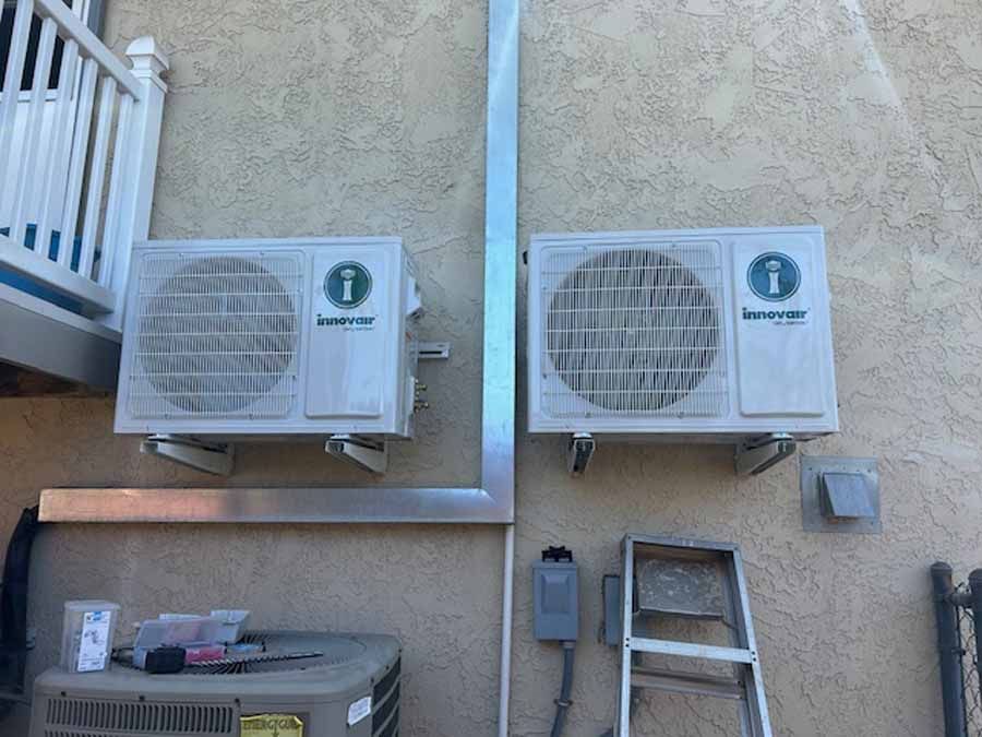 Two white wall-mounted air conditioners on a beige exterior wall with a ladder below.