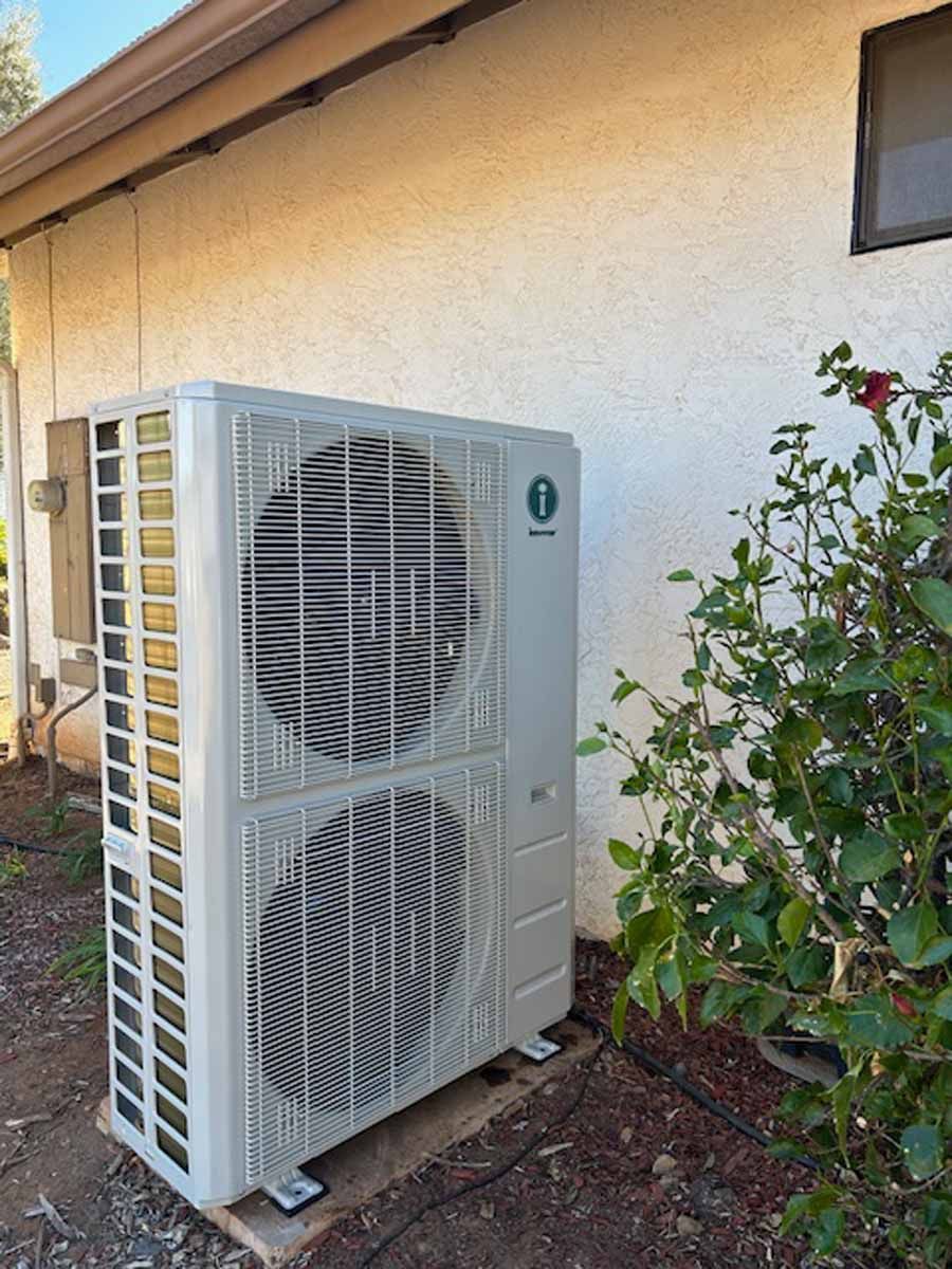Outdoor HVAC heat pump unit beside a stucco house wall and garden plants