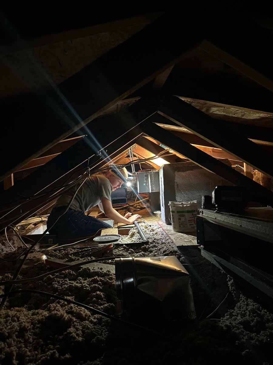 Person in a dark attic crawls under beams, working with tools near exposed insulation and a bright work light.