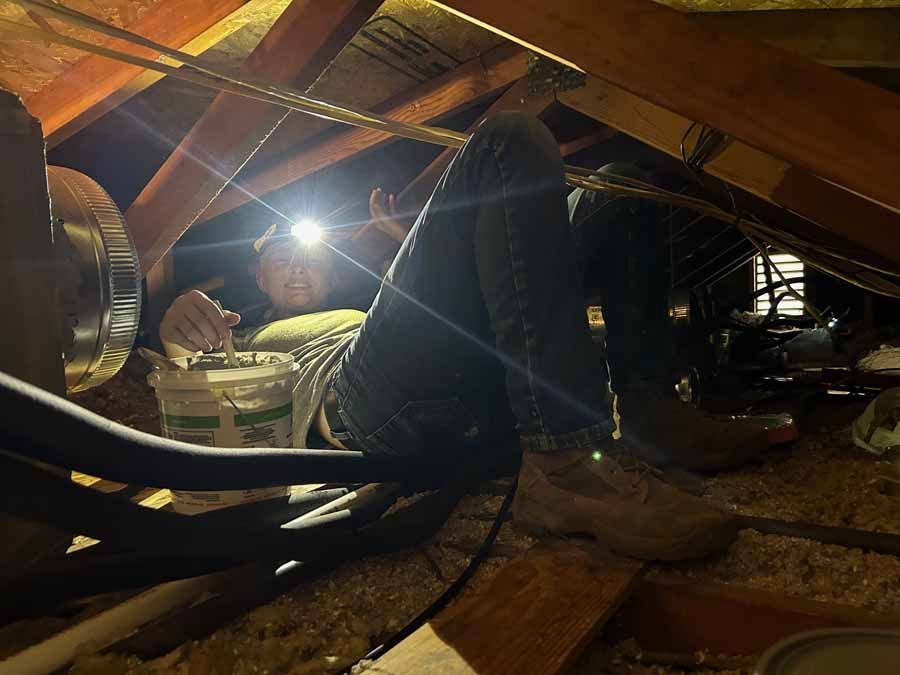 Two workers crouch in a dim attic with exposed wooden beams and a bright work light.