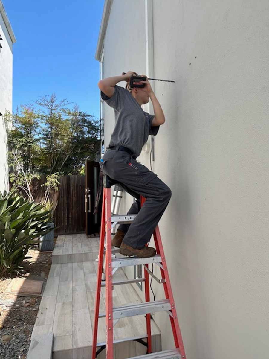 Man standing on a red ladder beside a white house, reaching up to work near the roofline.
