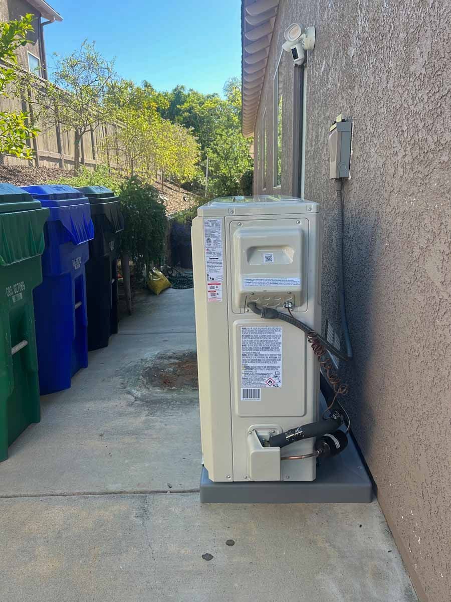 Outdoor air conditioning unit beside a stucco wall in a narrow alley, next to trash bins.
