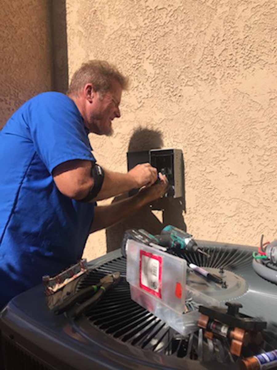Man in blue shirt repairing an outdoor wall-mounted electrical box next to HVAC equipment