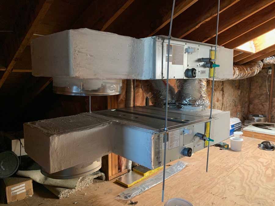 Large metal HVAC ducting and air handler installed in an attic with exposed wooden rafters.