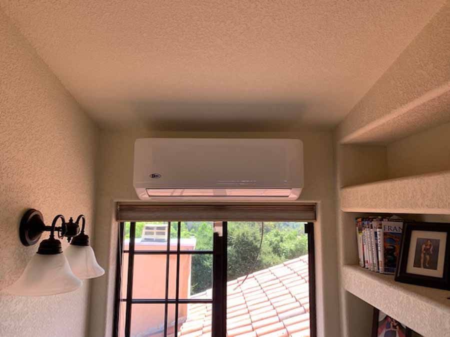 Bedroom with wall-mounted air conditioner above a window, framed photos on shelves, and a wall sconce.