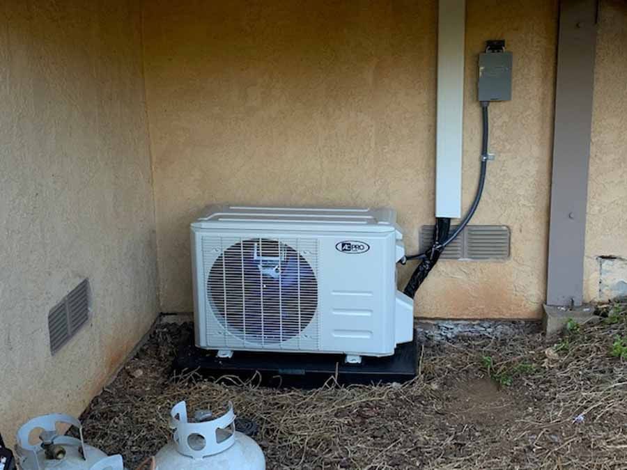 Outdoor air conditioning unit beside a beige house wall with electrical conduit and two discarded objects on the ground