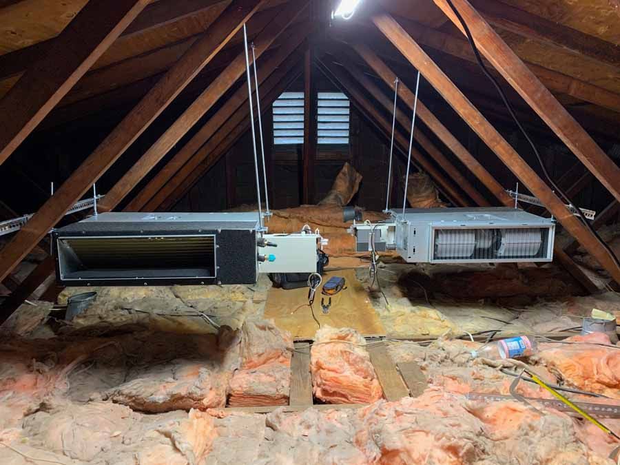 Attic with exposed wooden beams and two hanging HVAC ducts above insulation and brick supports
