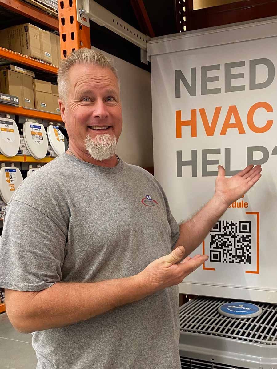 Man smiling and gesturing toward a “Need HVAC Help?” sign with a QR code in a warehouse aisle