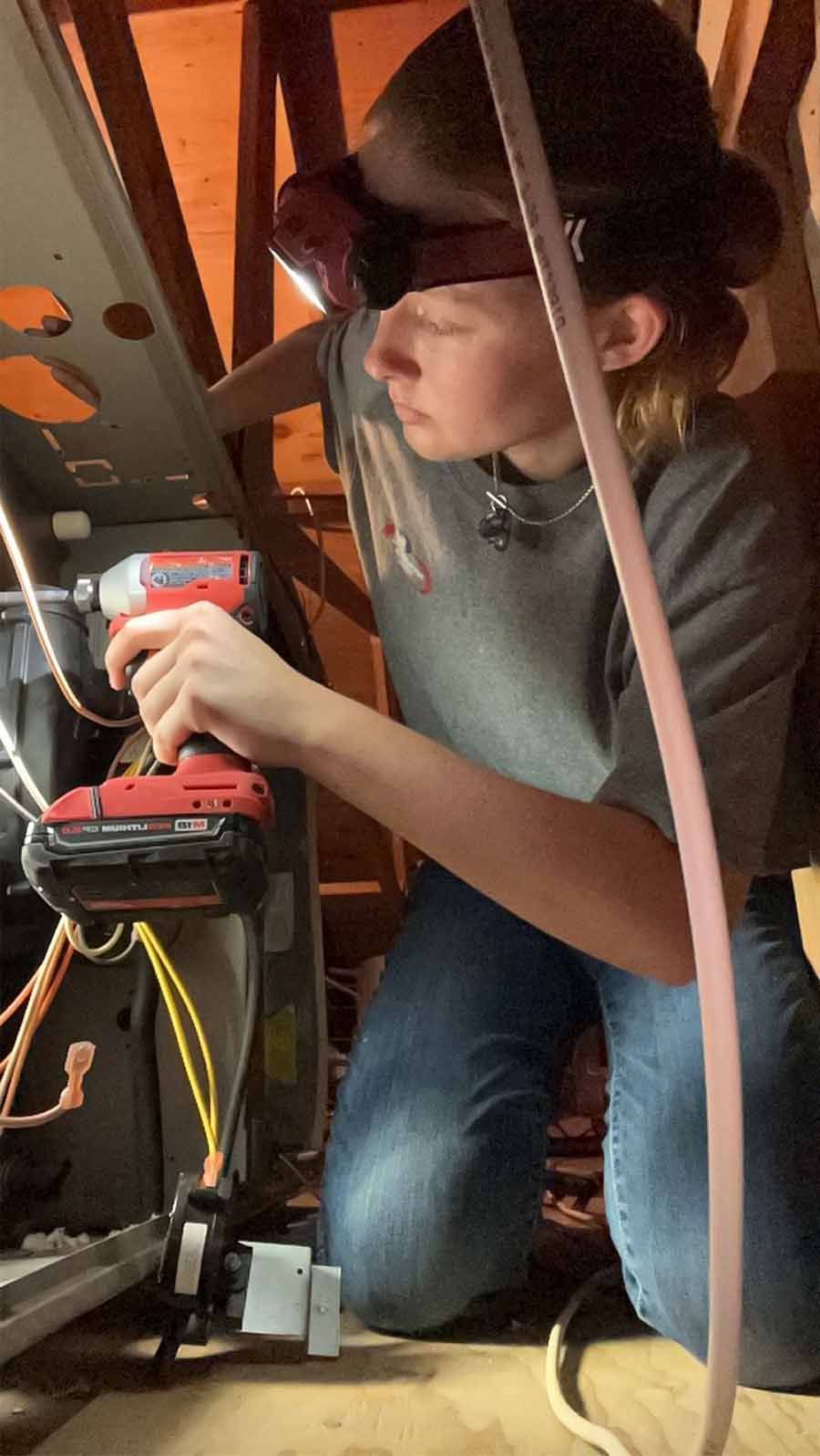 Person wearing a helmet operates a red power drill while kneeling inside a wooden structure.