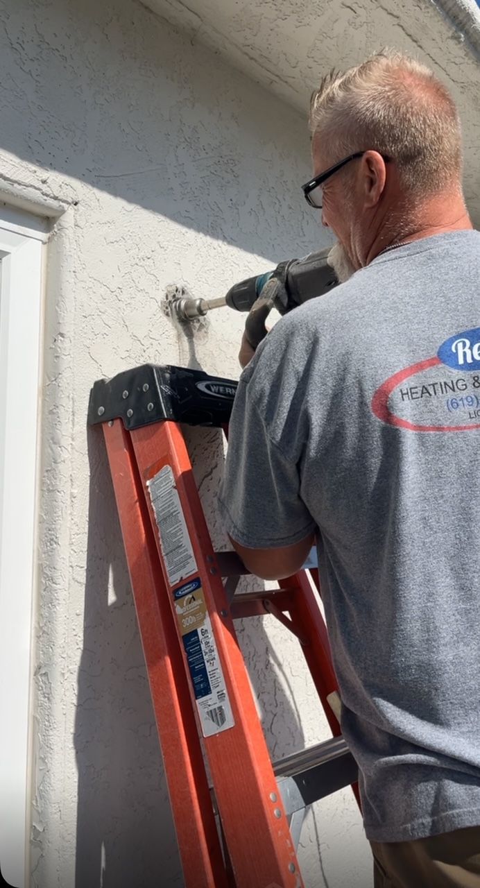 Man on an orange ladder drilling into a white exterior wall near a window