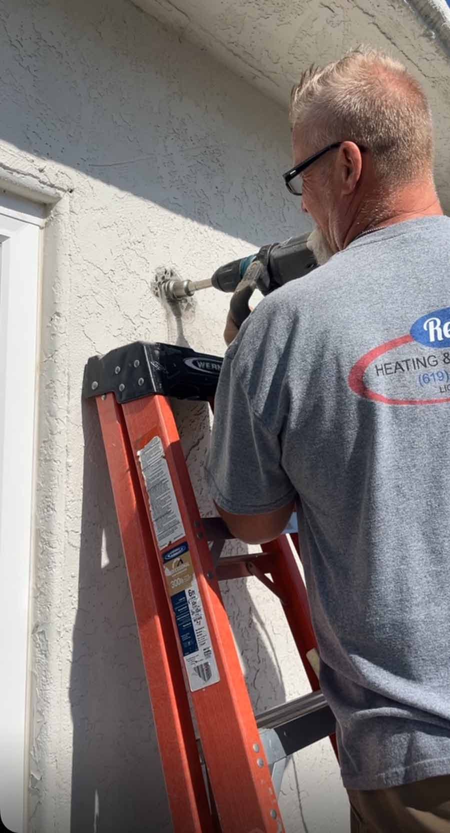 Worker using a drill on an exterior wall beside an orange ladder