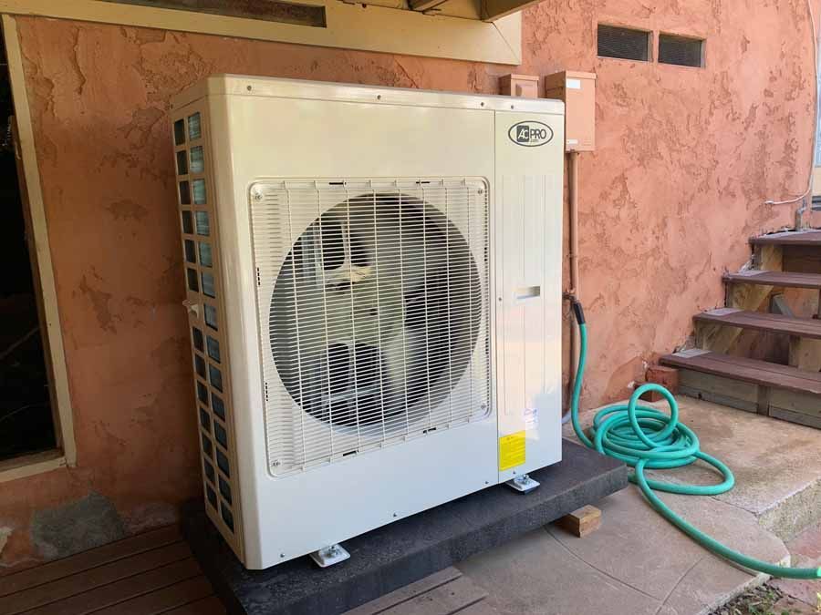 Outdoor air conditioning unit beside a pink stucco wall, with a green hose coiled nearby.
