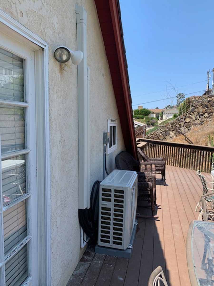 Wooden deck beside a white house with an air conditioner, chair, and hillside view under a blue sky
