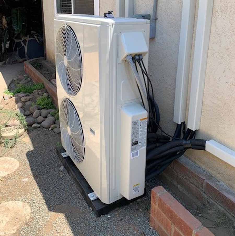 Outdoor air conditioner unit beside a house wall, with two large fans and visible wiring.