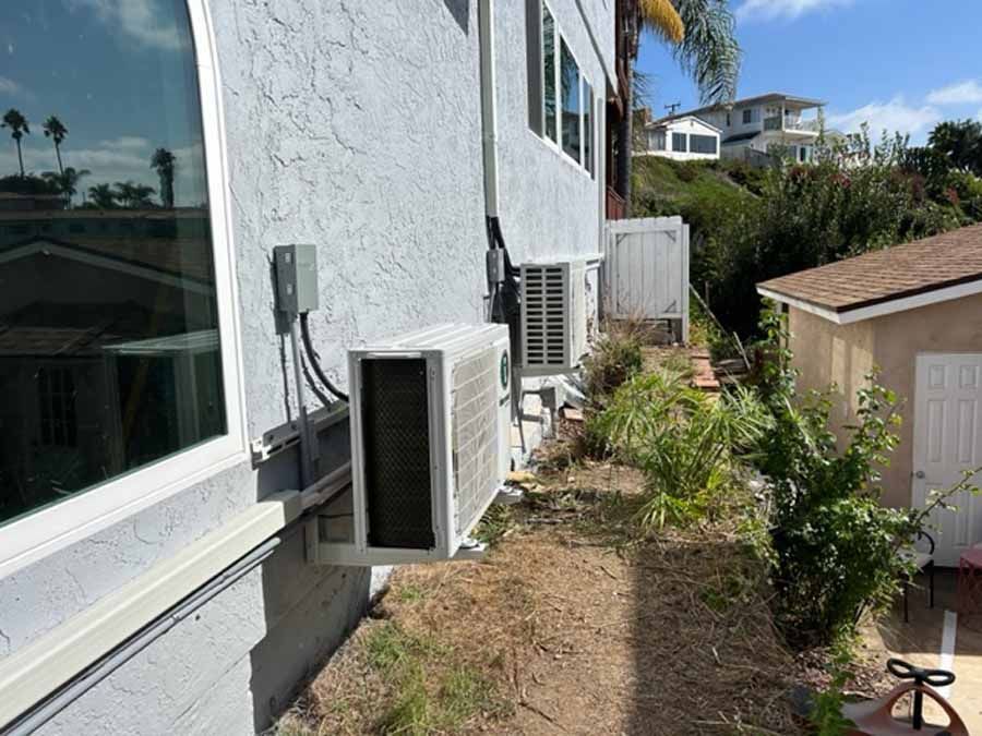 Side view of a house with two wall-mounted air conditioners beside a narrow dirt path and yard