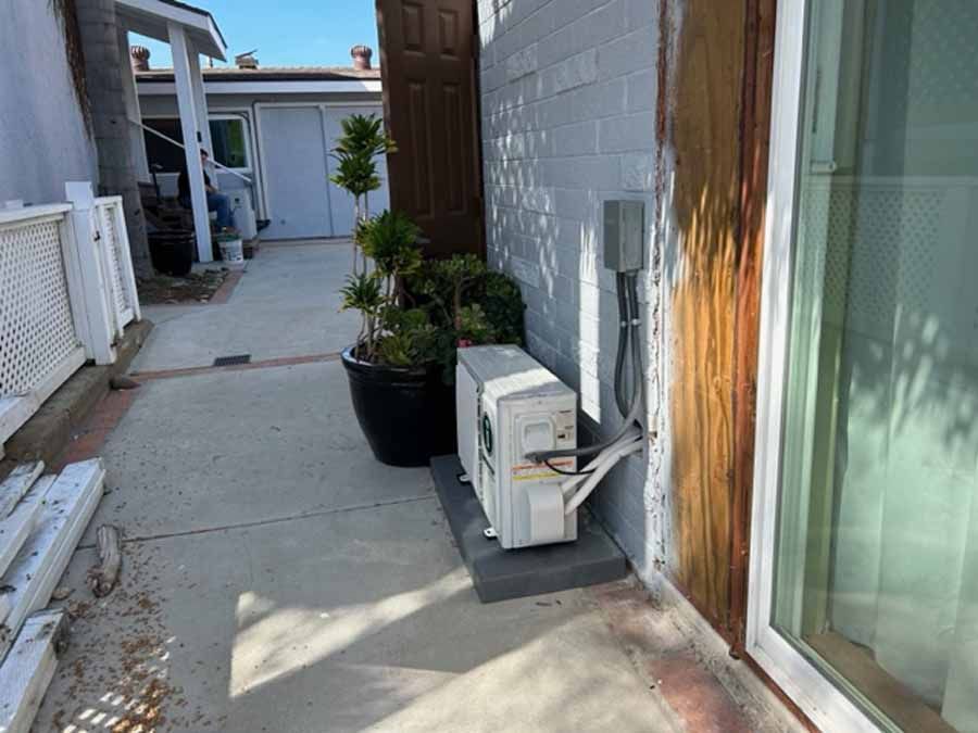 Narrow outdoor walkway beside a house with an air conditioner unit and potted plants along the wall.