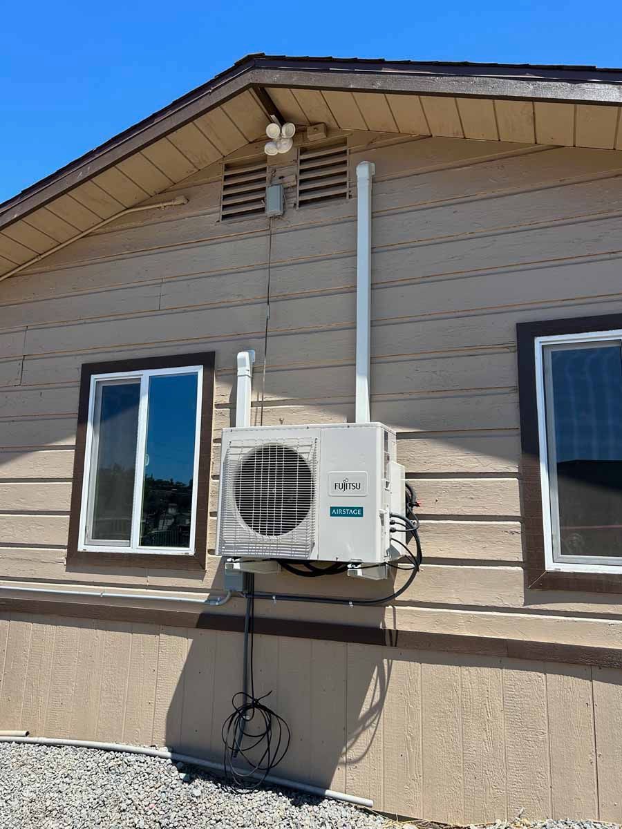 Exterior of a beige house with a wall-mounted air conditioner and white downspout under a blue sky