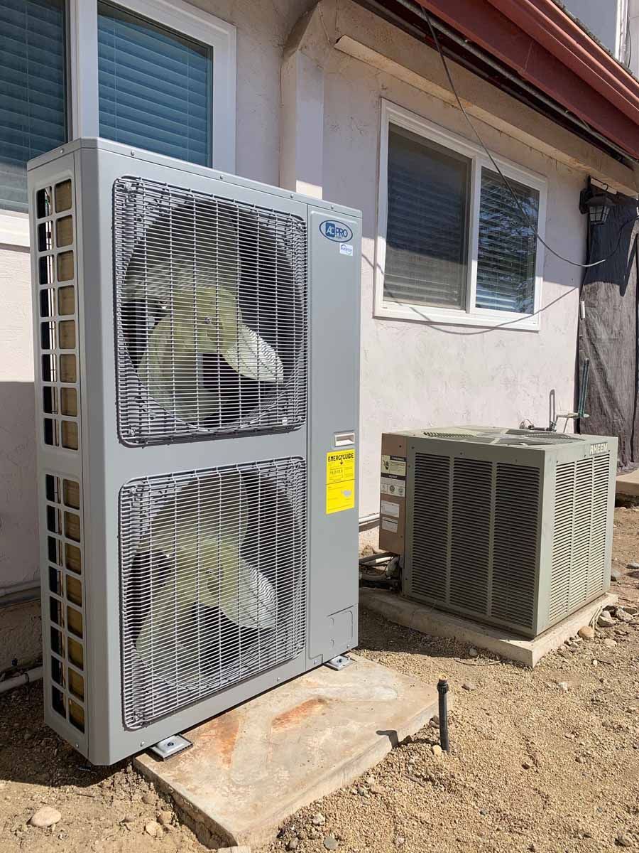 Outdoor HVAC units beside a house, including a large gray fan unit and a smaller beige condenser.