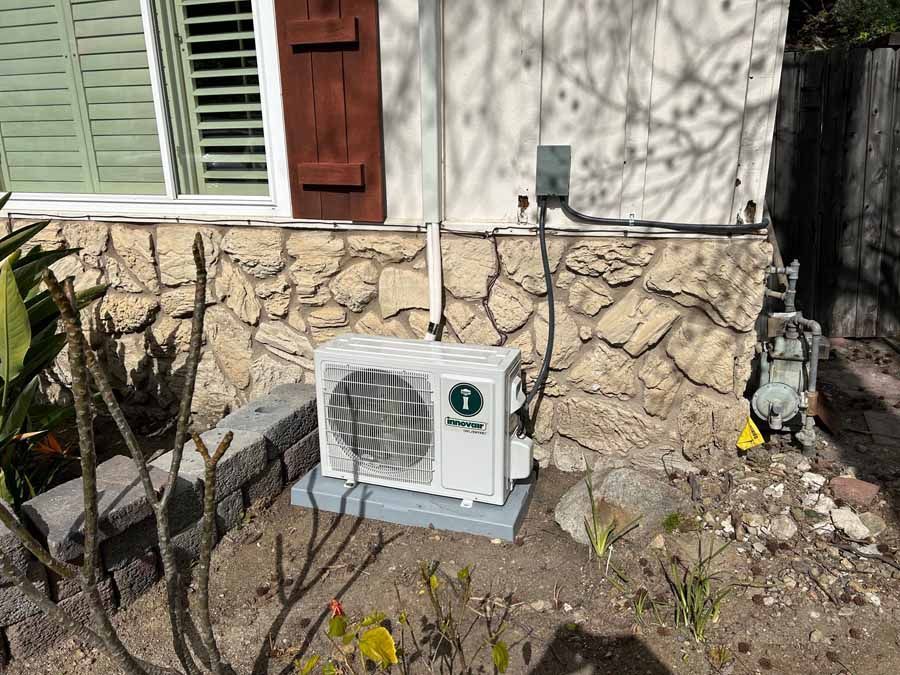 Outdoor air conditioner unit beside a stone house wall with plants in front