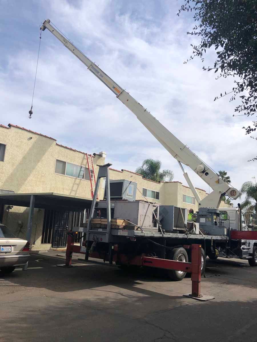Crane truck lifting equipment beside a building, with parked cars and a cloudy sky.