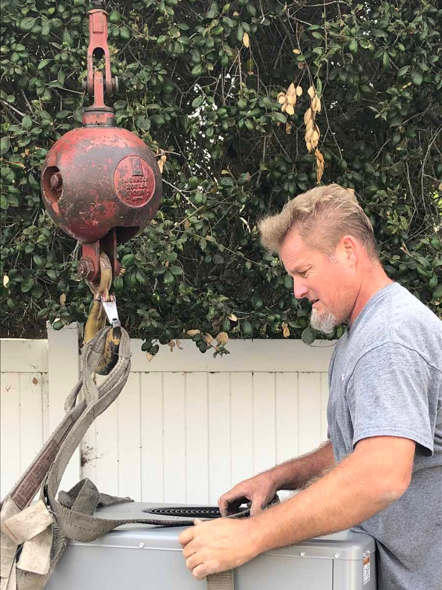Man operating a red hoist to lift a gray kayak in a backyard.