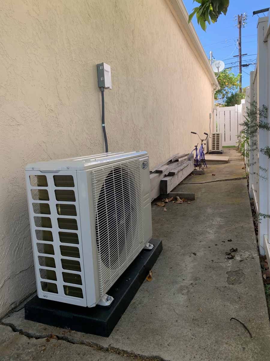 Outdoor heat pump units beside a beige wall in a narrow concrete alley.