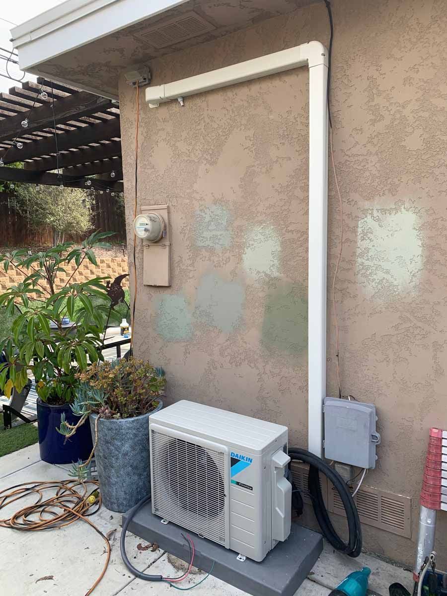 Outdoor wall with an air conditioner, pipes, electrical box, and potted plants beside a patio.