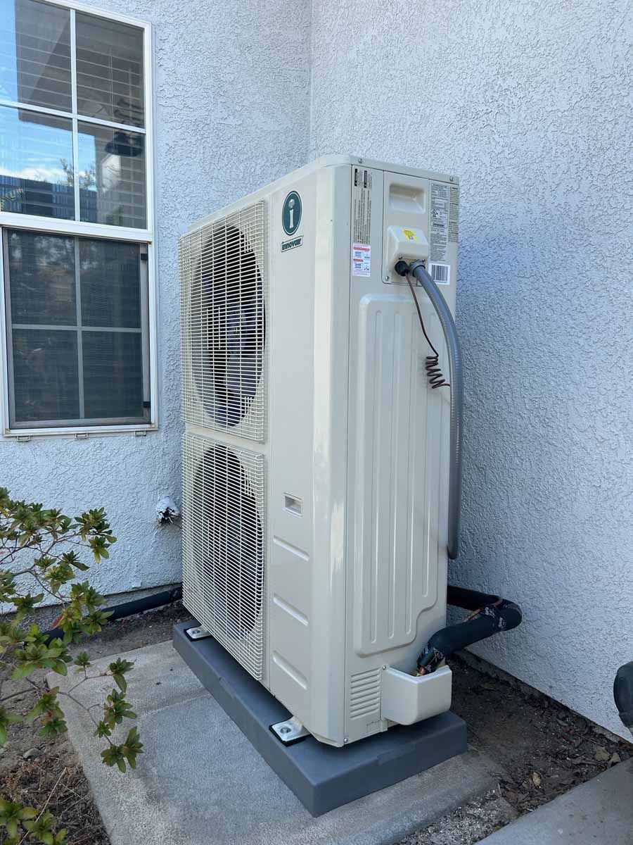 Outdoor air conditioning unit beside a stucco house wall near a window and plants