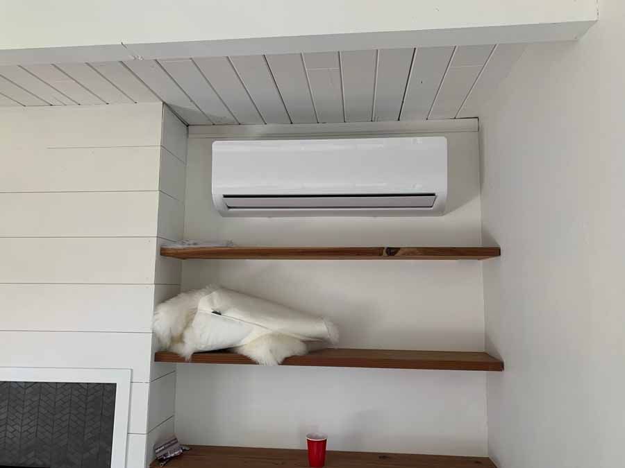 White cat sleeping on a wooden shelf beneath a wall-mounted air conditioner.