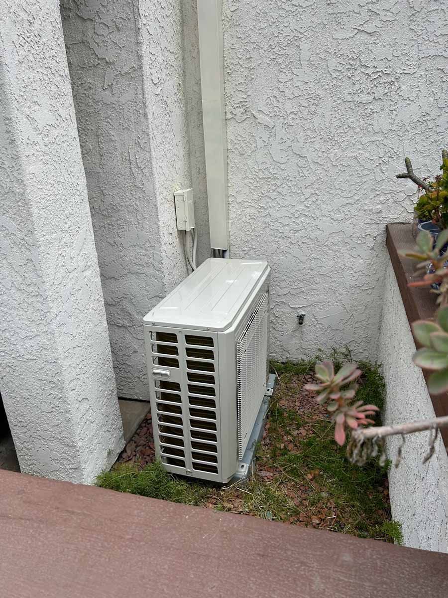 Outdoor air conditioner unit beside a stucco wall and potted plants on a narrow patio