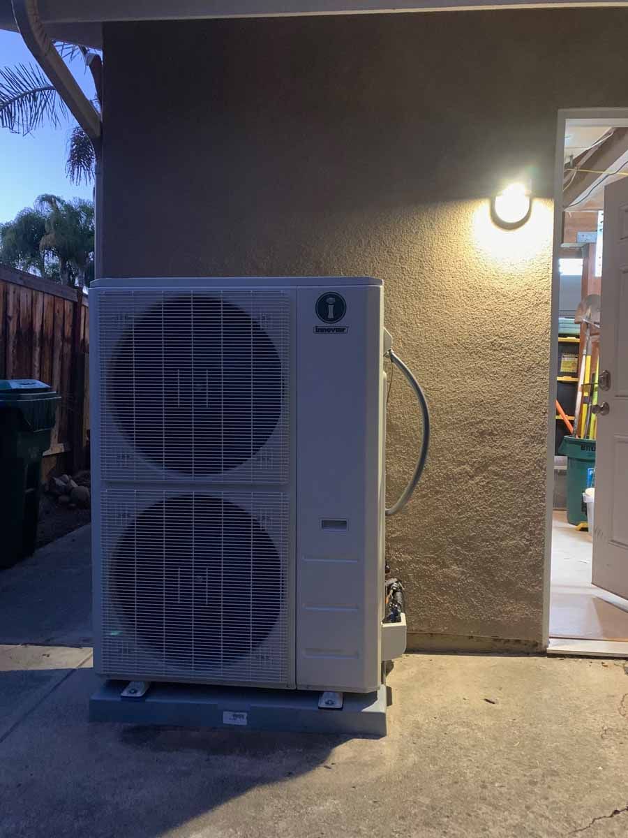 Outdoor air-conditioning unit beside a house at dusk, under a lit exterior wall lamp.