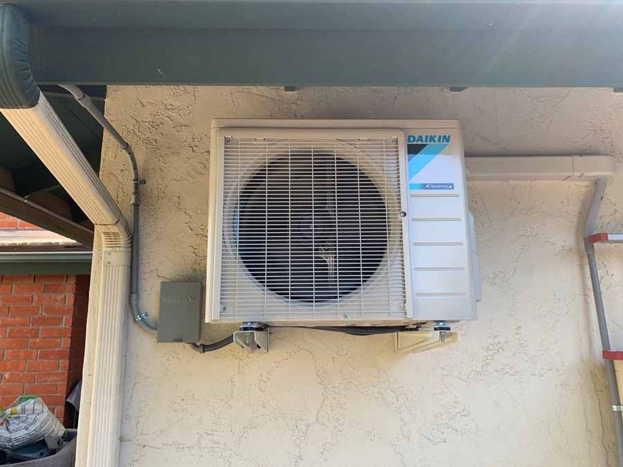 Wall-mounted air conditioner outdoor unit on a beige stucco exterior wall under a roof overhang.