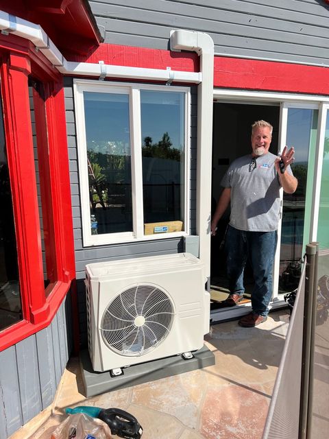 Man standing on a rooftop deck beside a window and air conditioner, giving a thumbs-up.